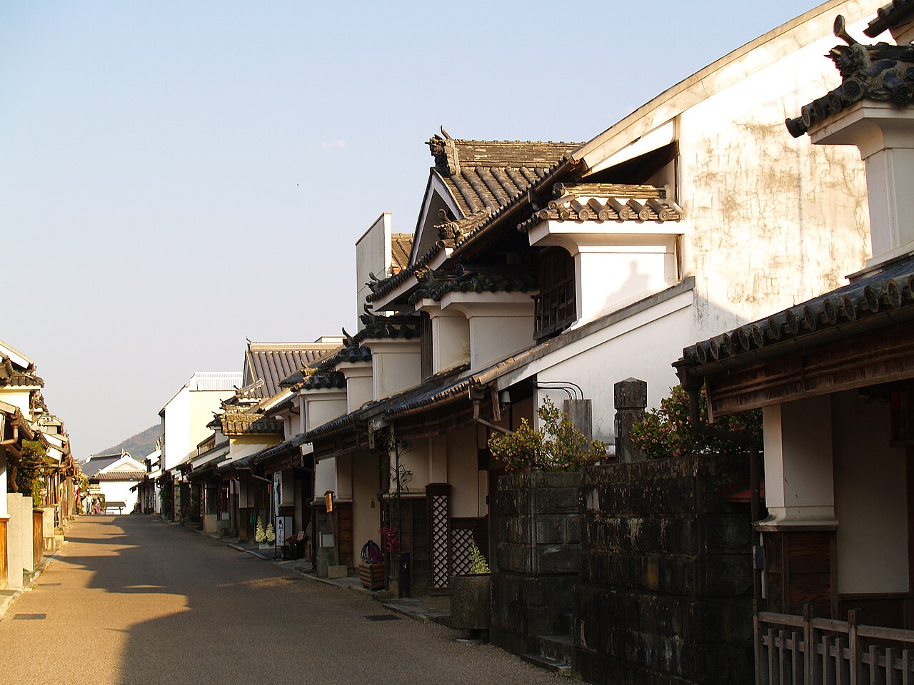 A row of whitewashed udatsu houses with tiled roofs along a quiet paved street in Wakimachi, lit by low warm sunlight with hills visible in the distance.