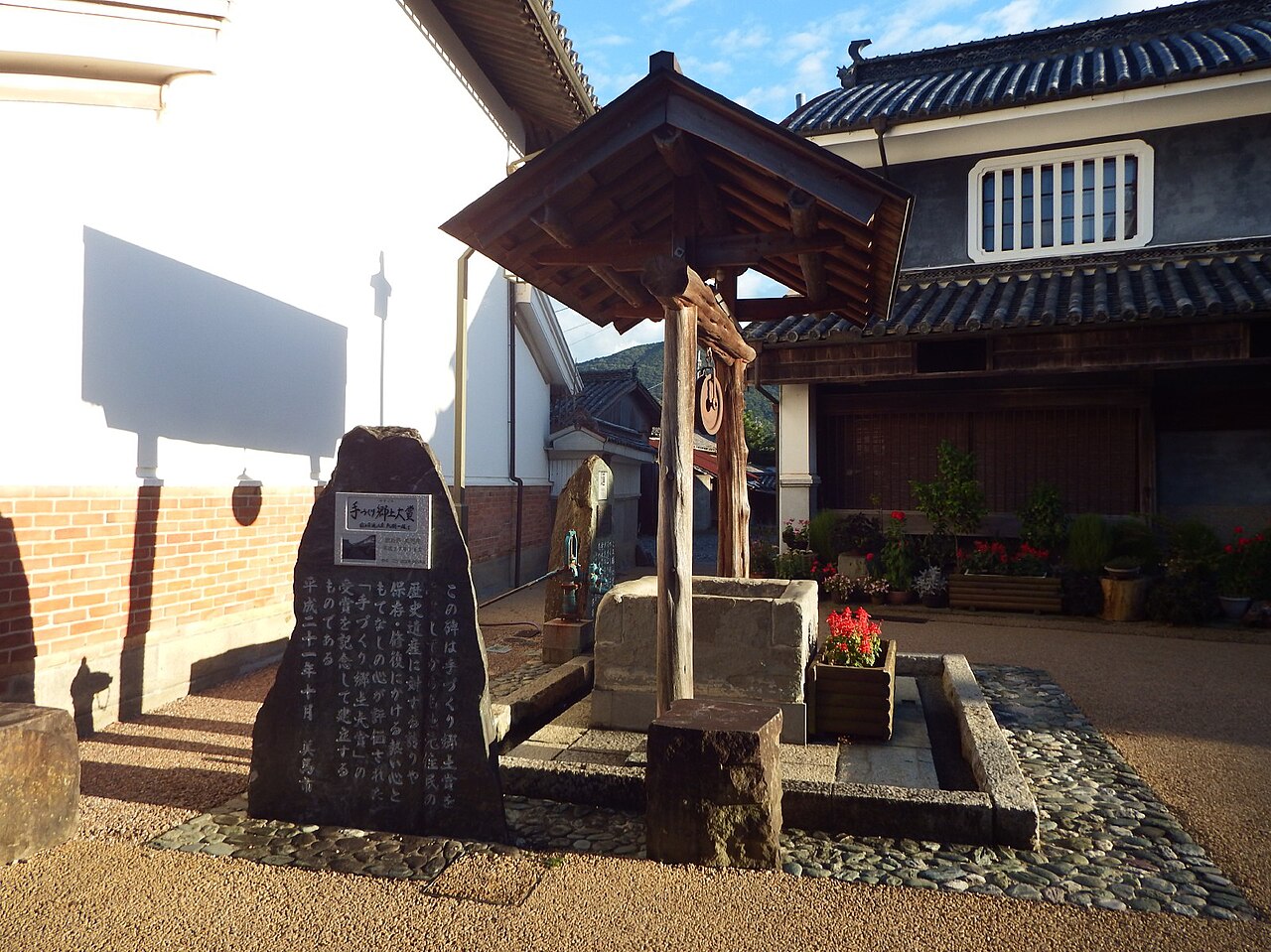 A covered stone well with a wooden roof and a stone monument inscribed with Japanese text, standing in a courtyard in front of an udatsu-walled building in Wakimachi.