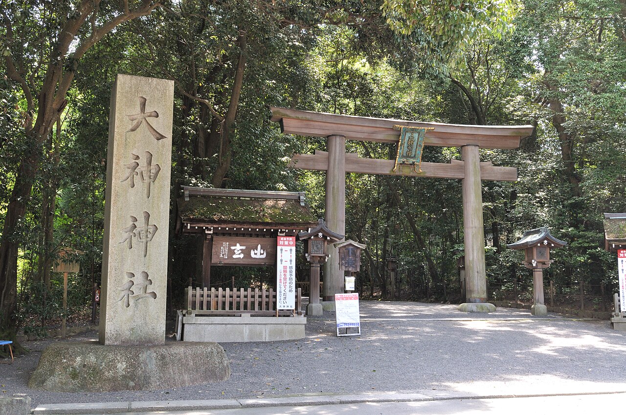 Stone torii gate on the approach to Omiwa Shrine, with a tall stone pillar carved with the shrine name in the foreground and a few visitors under the trees