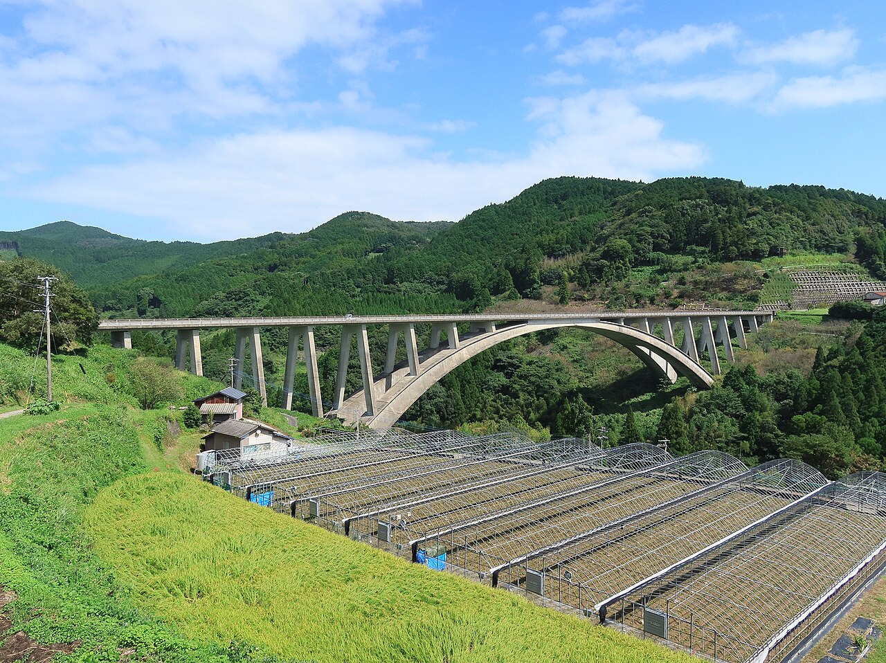 A long concrete arch bridge crossing a green valley in the Joyo area of Yame, with covered farm fields and terraced slopes in the foreground
