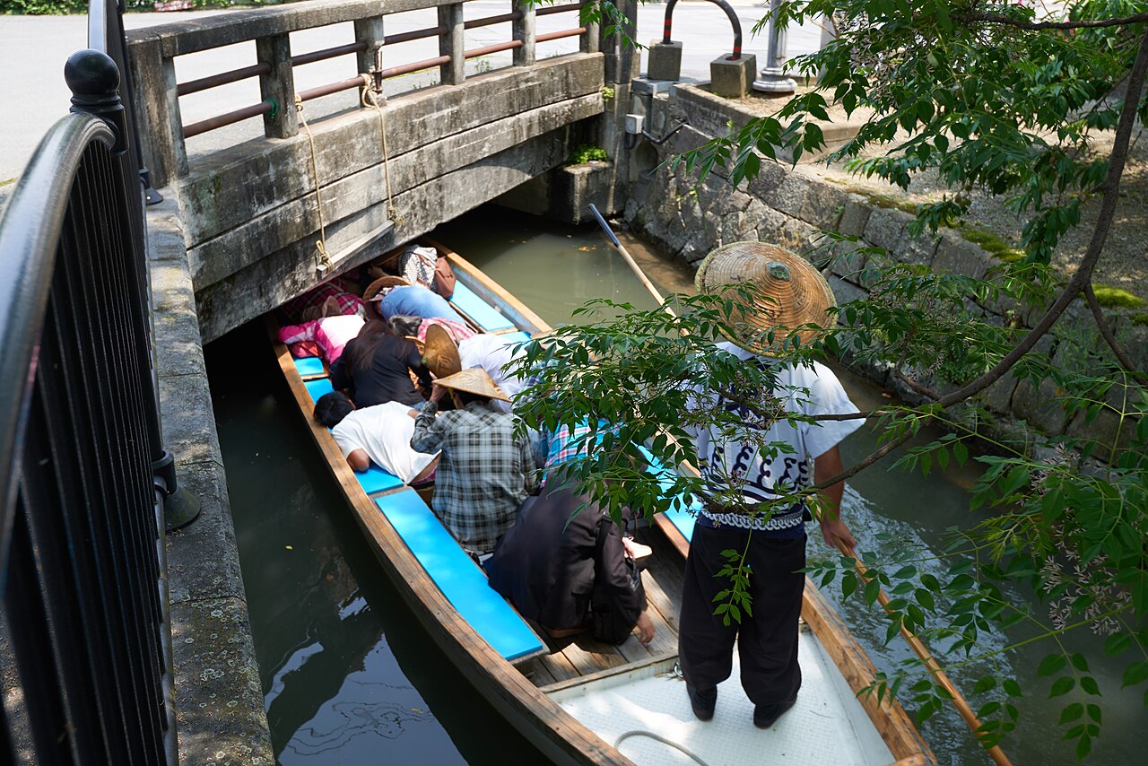Passengers bend low in a river cruise boat as it passes under a low stone bridge in Yanagawa