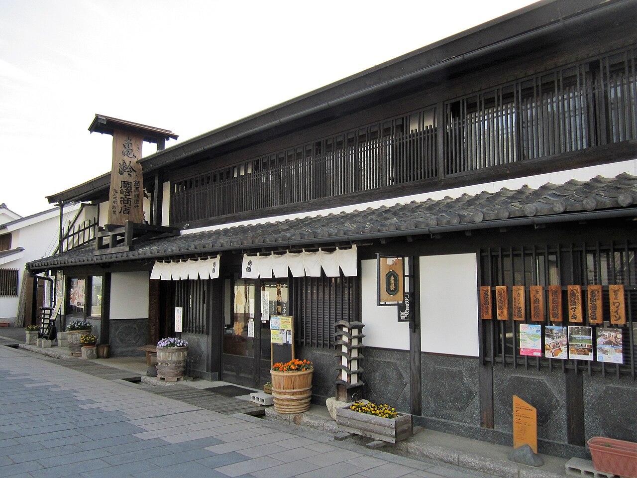 A long dark lattice-fronted sake shop on Yanagimachi with a wooden signboard, noren curtains and barrels of flowers