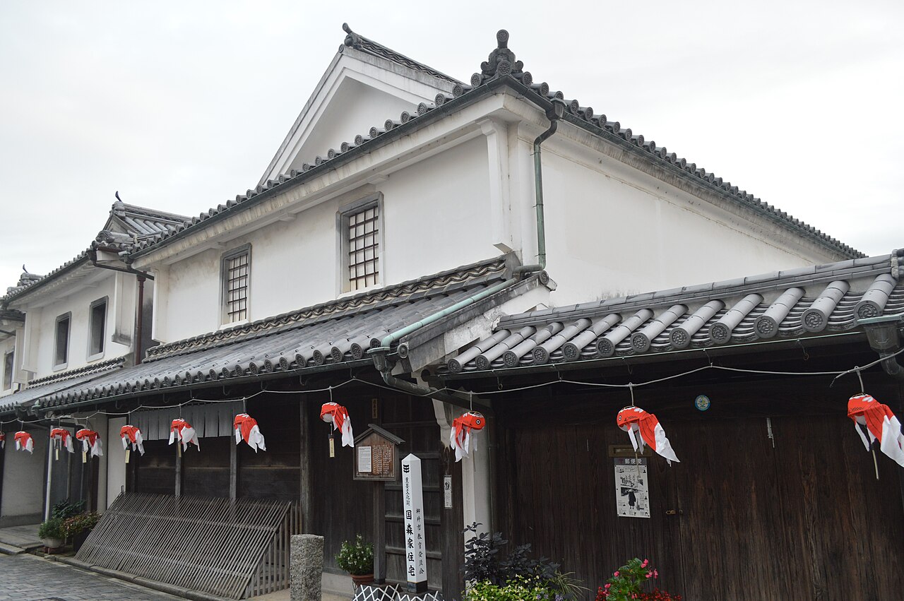 White-plastered storehouse of the Kunimori House with a tiled roof, a stone marker and a row of red-and-white goldfish lanterns hanging under the dark timber eaves