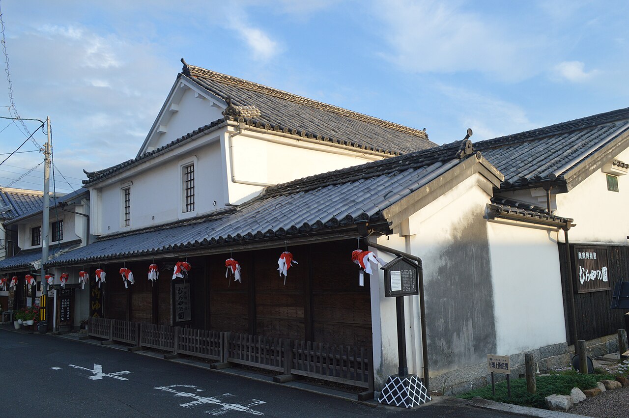 Two-storey white-walled merchant house museum Muroya-no-sono seen from the road at dusk, with goldfish lanterns hanging along its dark wooden lower wall