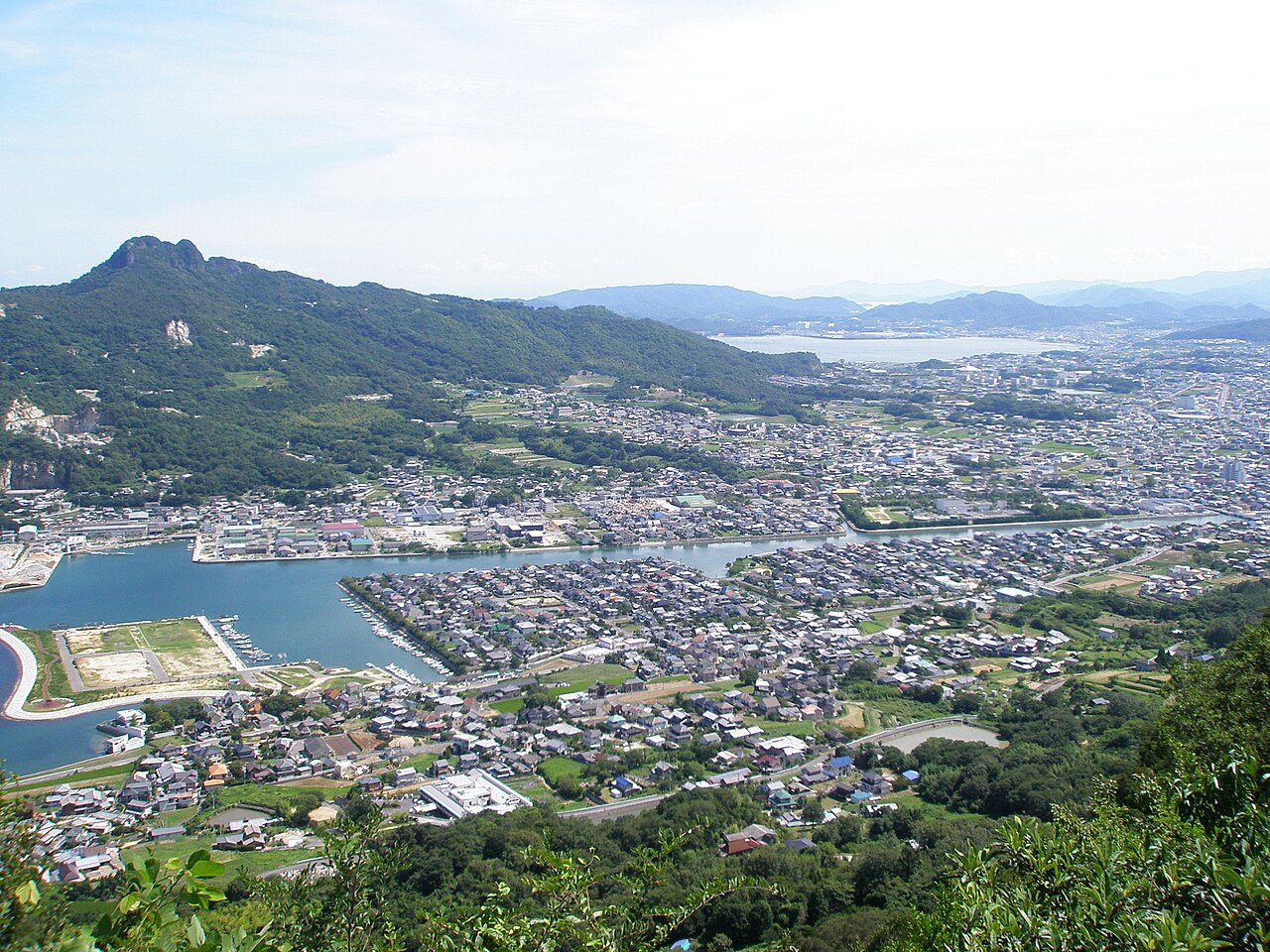 A wide view from a hillside viewpoint over a coastal town and a bay, with a rocky mountain ridge rising on the left