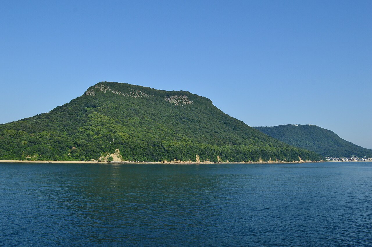 The flat-topped, green-forested bulk of Mount Yashima seen across calm blue water from the sea