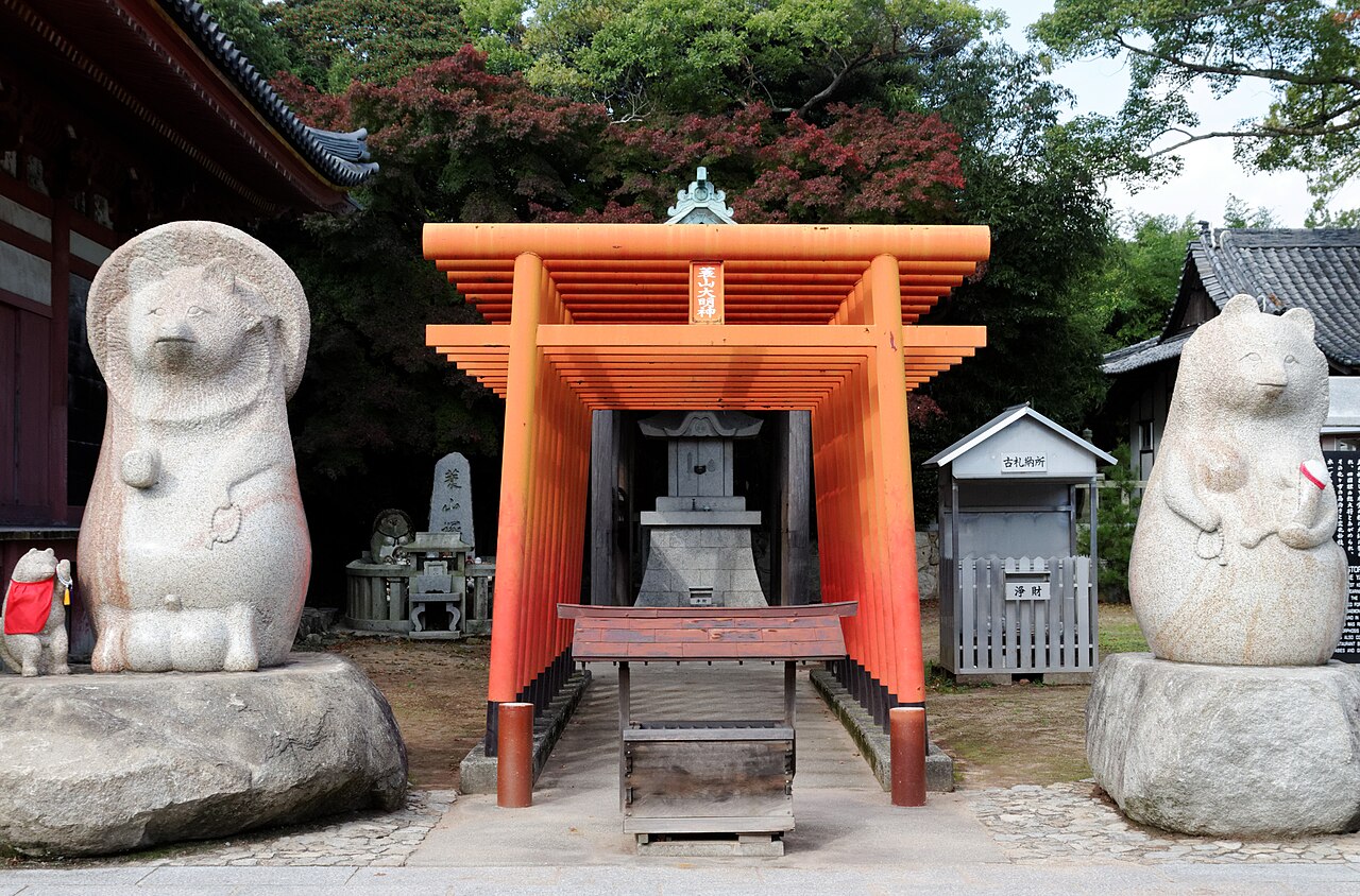 Two large white stone tanuki statues flanking a small orange torii and stone lantern in a temple courtyard