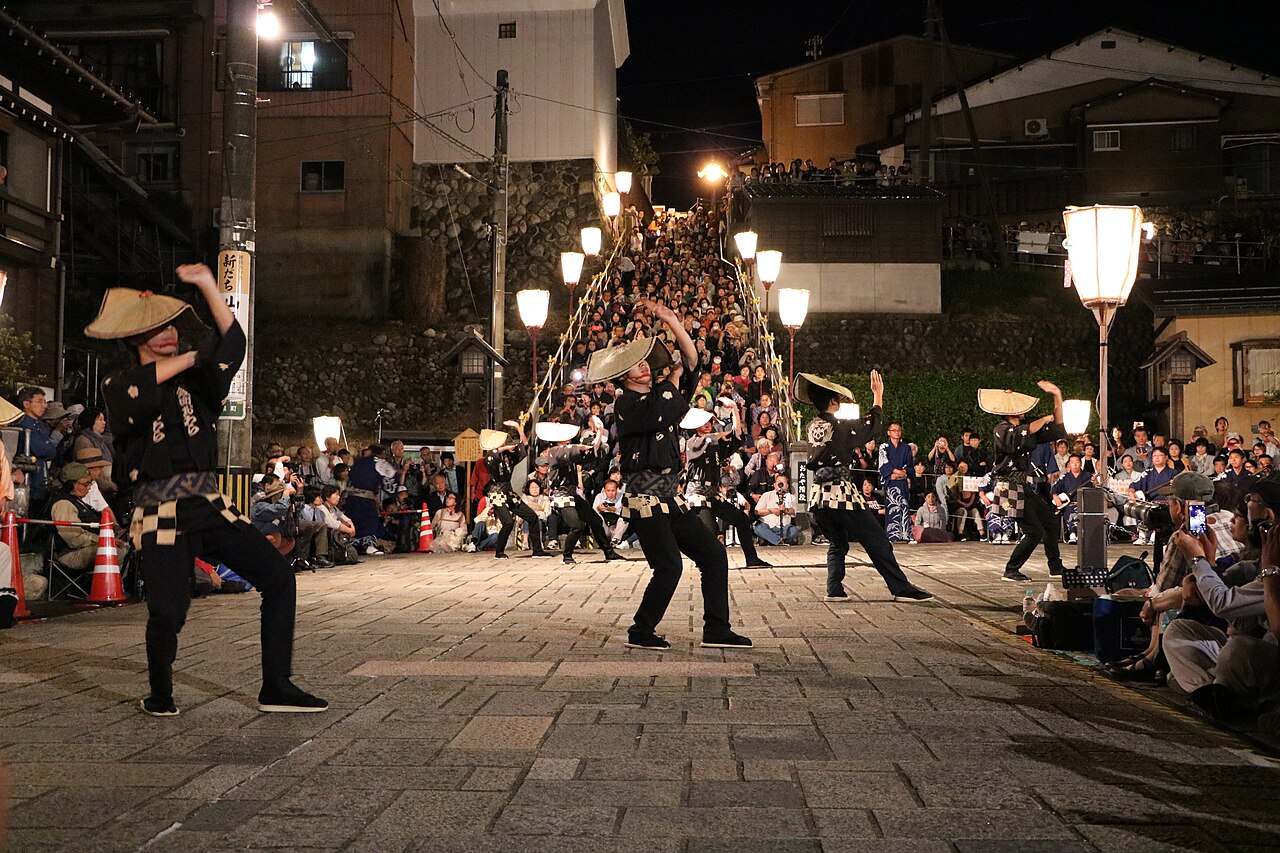 Night performance of Owara Kaze no Bon in Yatsuo: dancers in black costumes and straw hats on a paved street, with a crowd on the stone steps behind and lanterns glowing.
