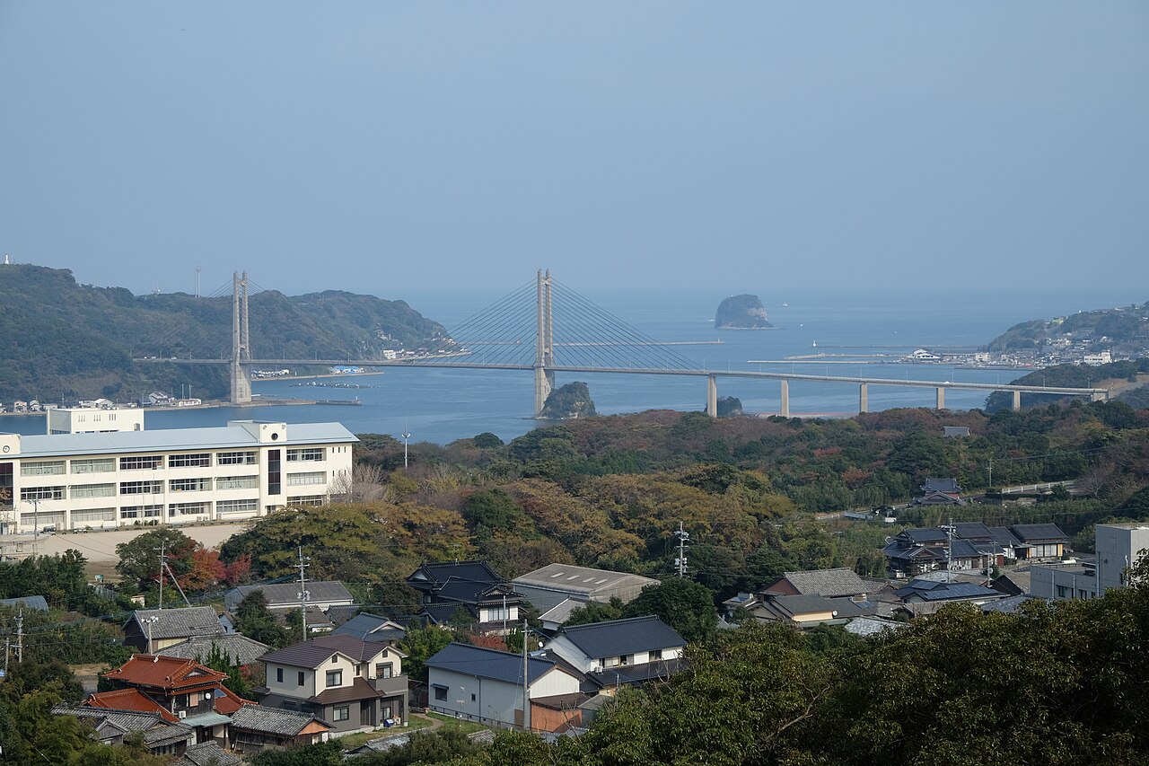 A cable-stayed bridge spans a blue bay seen from a hillside above a small village, with rocky islets in the water and autumn-tinted trees in the foreground