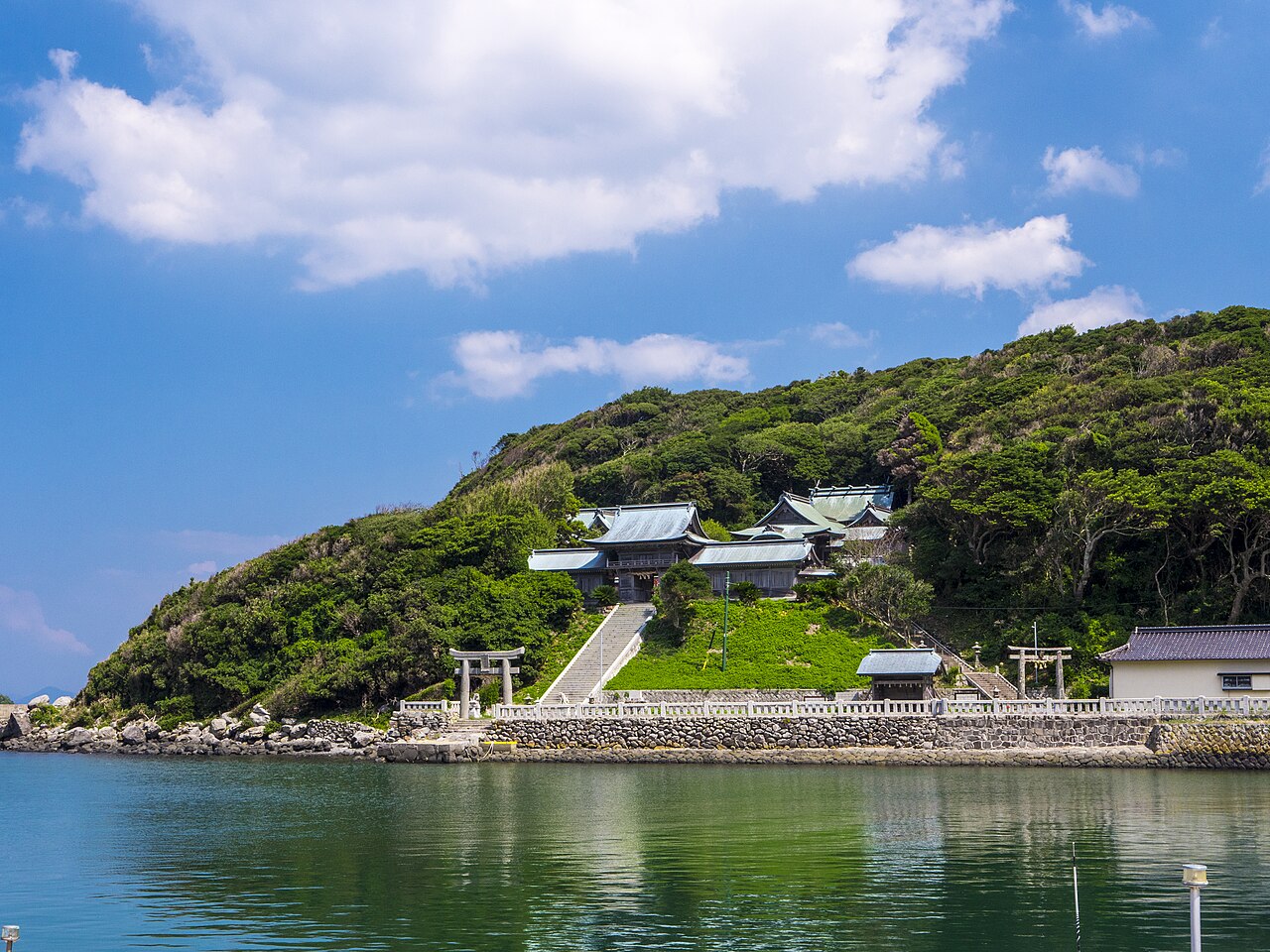 A shrine with torii gates and a stone stairway set against a green wooded hillside, seen across the water from the opposite shore