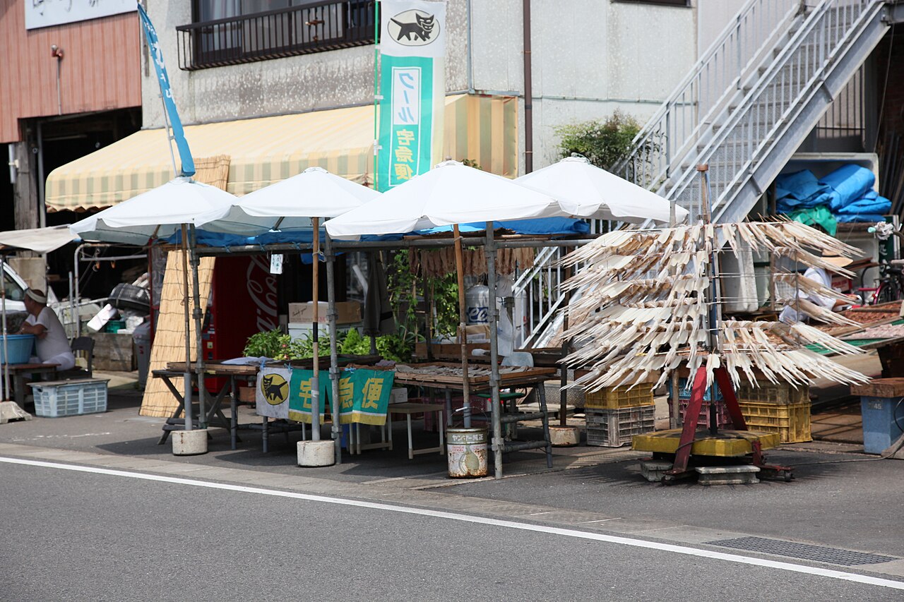 Roadside morning-market stalls under white umbrellas in Yobuko, with split squid hung out to dry on a rack at the right