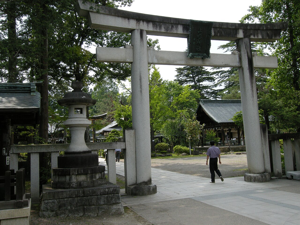 A weathered stone torii and a stone lantern at the entrance of Uesugi Shrine in Yonezawa, with a tiled shrine hall and green trees beyond.