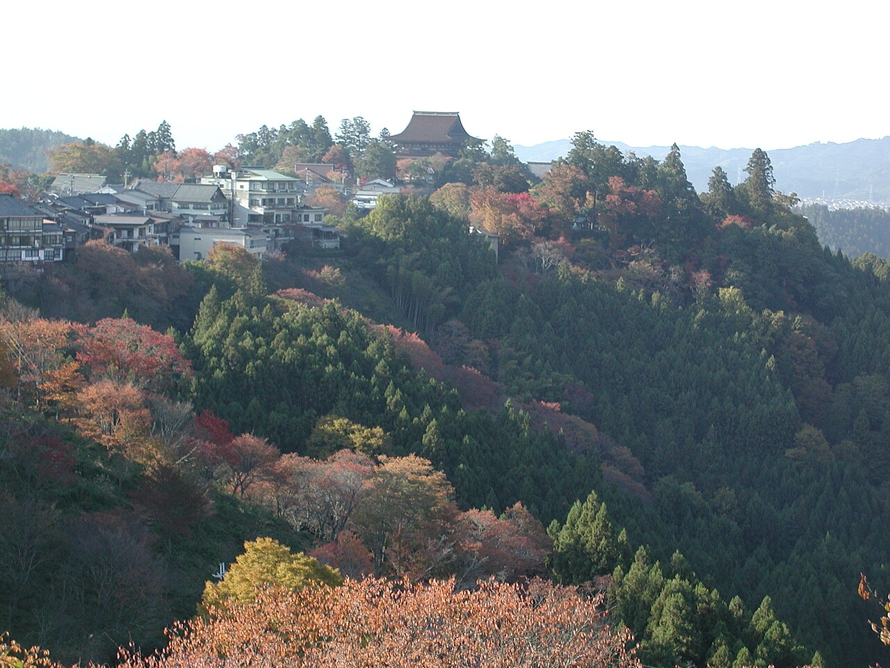 The hillside village of Yoshinoyama in autumn, with rooftops climbing a forested slope toward the distant Zao-do hall and mixed red and green foliage in the foreground