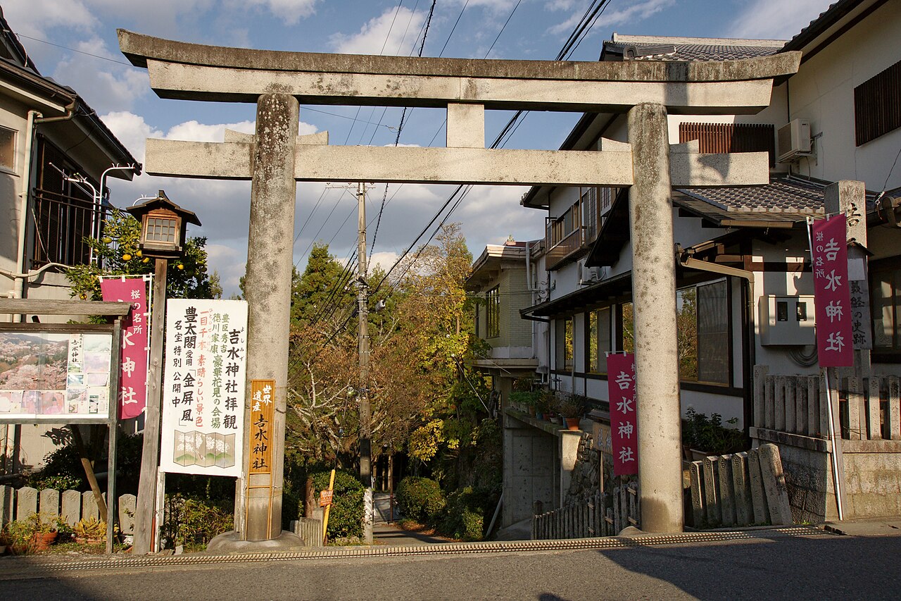 A weathered stone torii gate at the entrance to Yoshimizu Shrine on Mount Yoshino, with pink banners, a stone lantern and old wooden houses lining the narrow road beside it