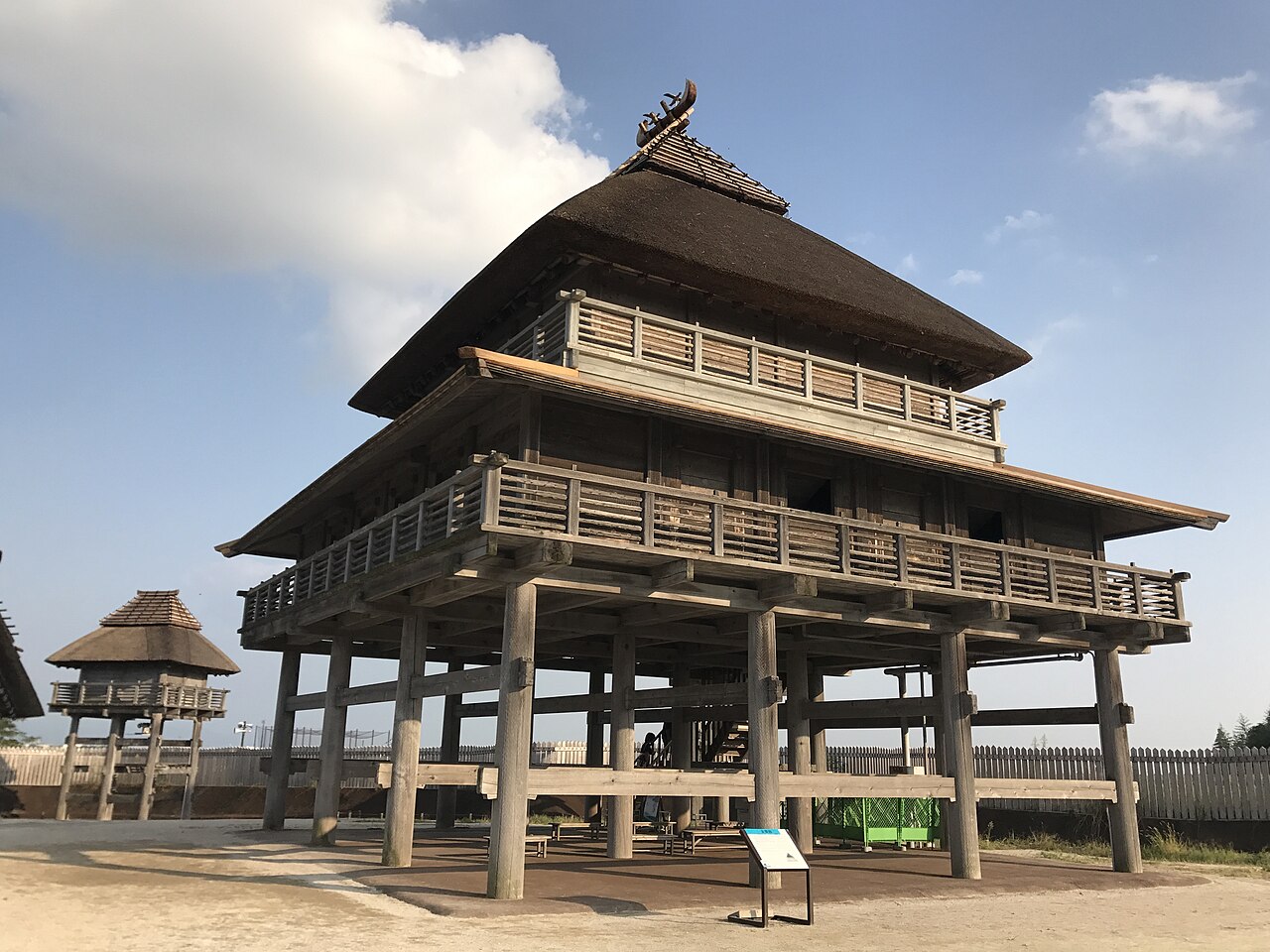 The reconstructed main hall of the northern inner enclosure at Yoshinogari, a tall raised wooden building with a layered thatched roof and a veranda under a blue sky.