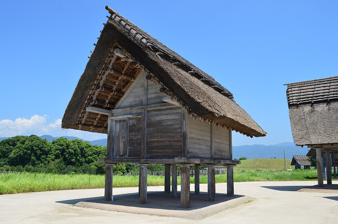 A reconstructed raised-floor storehouse at Yoshinogari with a steep thatched roof on short wooden posts, green fields and hills behind.