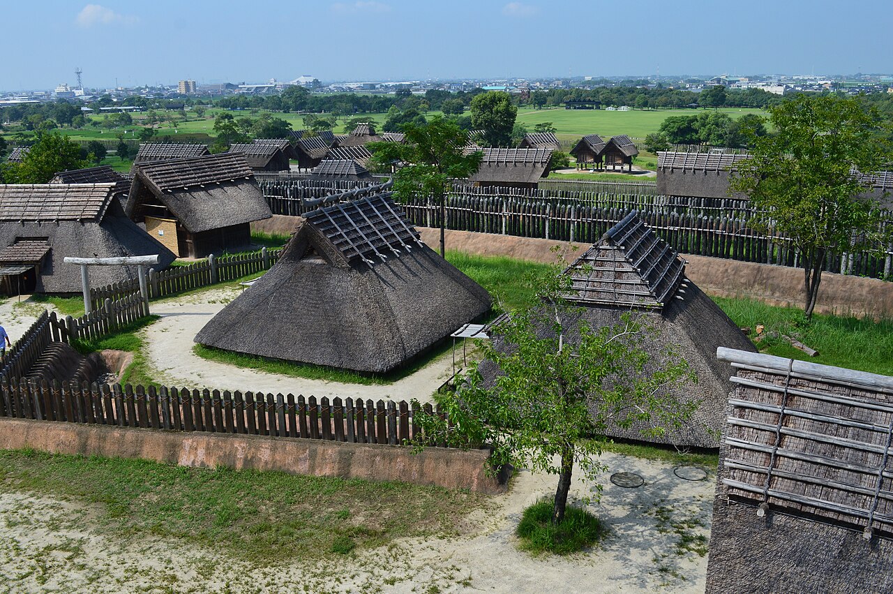 Looking down over the reconstructed thatched houses and wooden fences of the southern inner enclosure at Yoshinogari, with flat farmland stretching to the horizon.