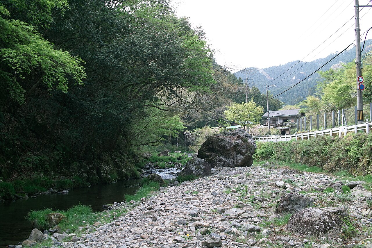 The stony bed of the Yozawa stream at Akiruno in western Tokyo, clear water running past boulders with village roofs and wooded ridges above.