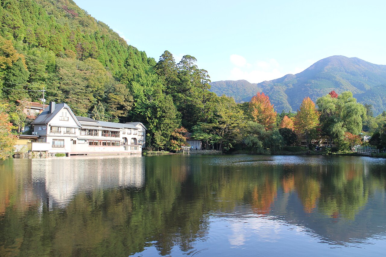 Lake Kinrin on a clear autumn morning, with a lakeside inn, a small torii and early red maples reflected in still water below green hills