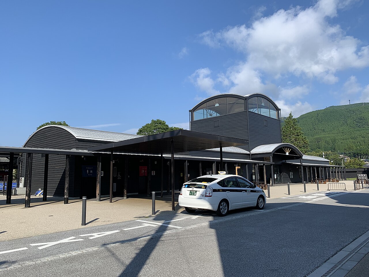 The black timber Yufuin Station building with its arched roofs and glass lantern tower, a taxi waiting in front