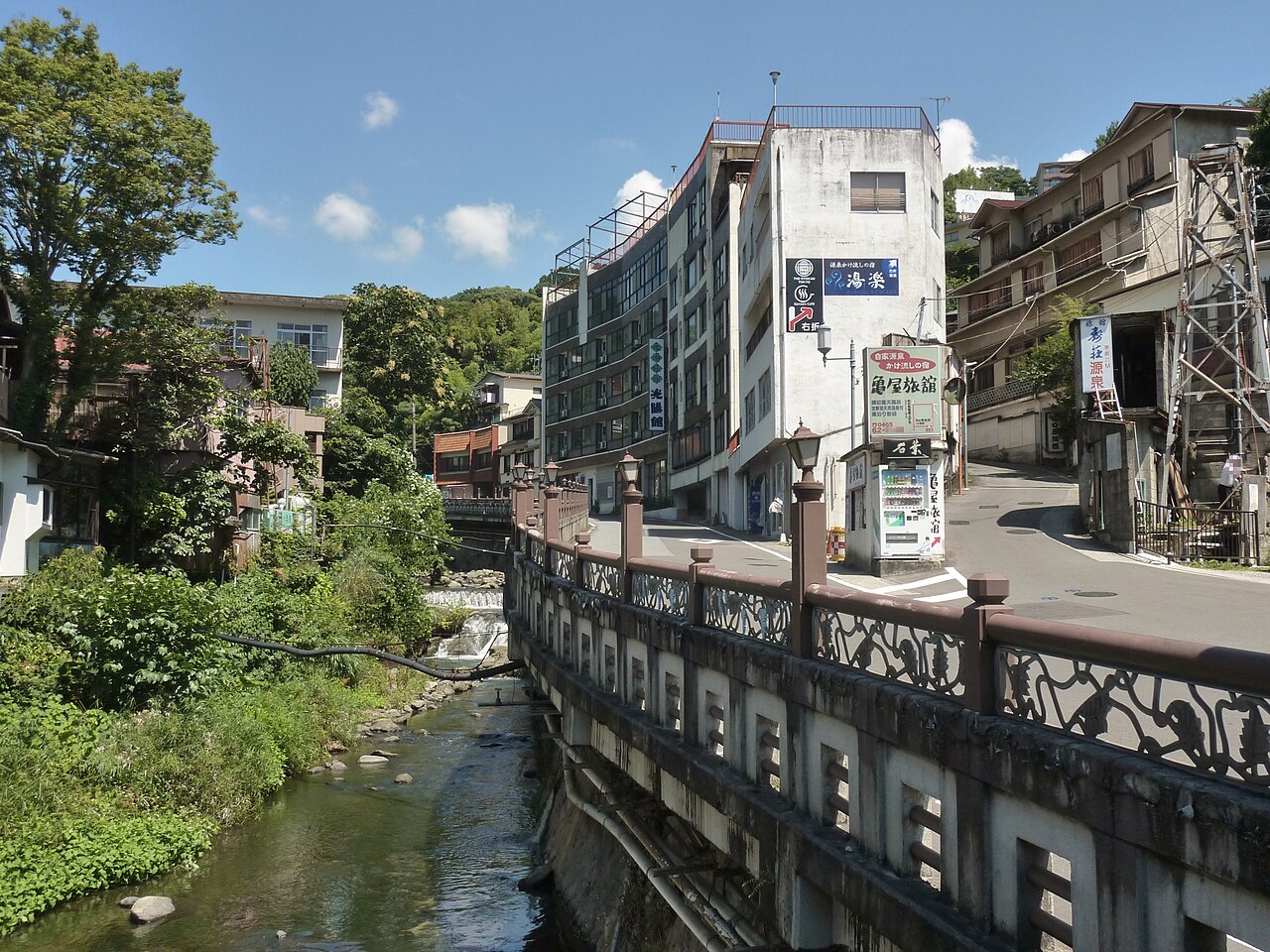A shallow stream running through a hillside town between concrete embankments, with multi-storey inns and a railed road along the bank under a blue sky.