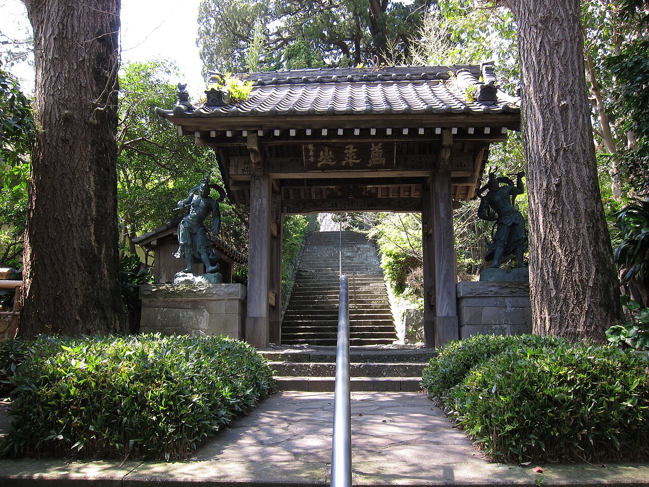 A tiled-roof temple gate with two guardian statues beside it, seen from the foot of a stone stairway shaded by tall cedar trees.