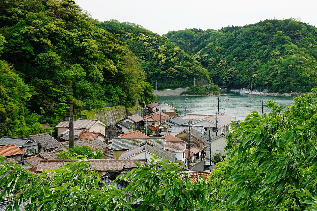 Orange-tiled rooftops of a small harbor settlement seen from above, with a calm inlet and forested hills beyond.