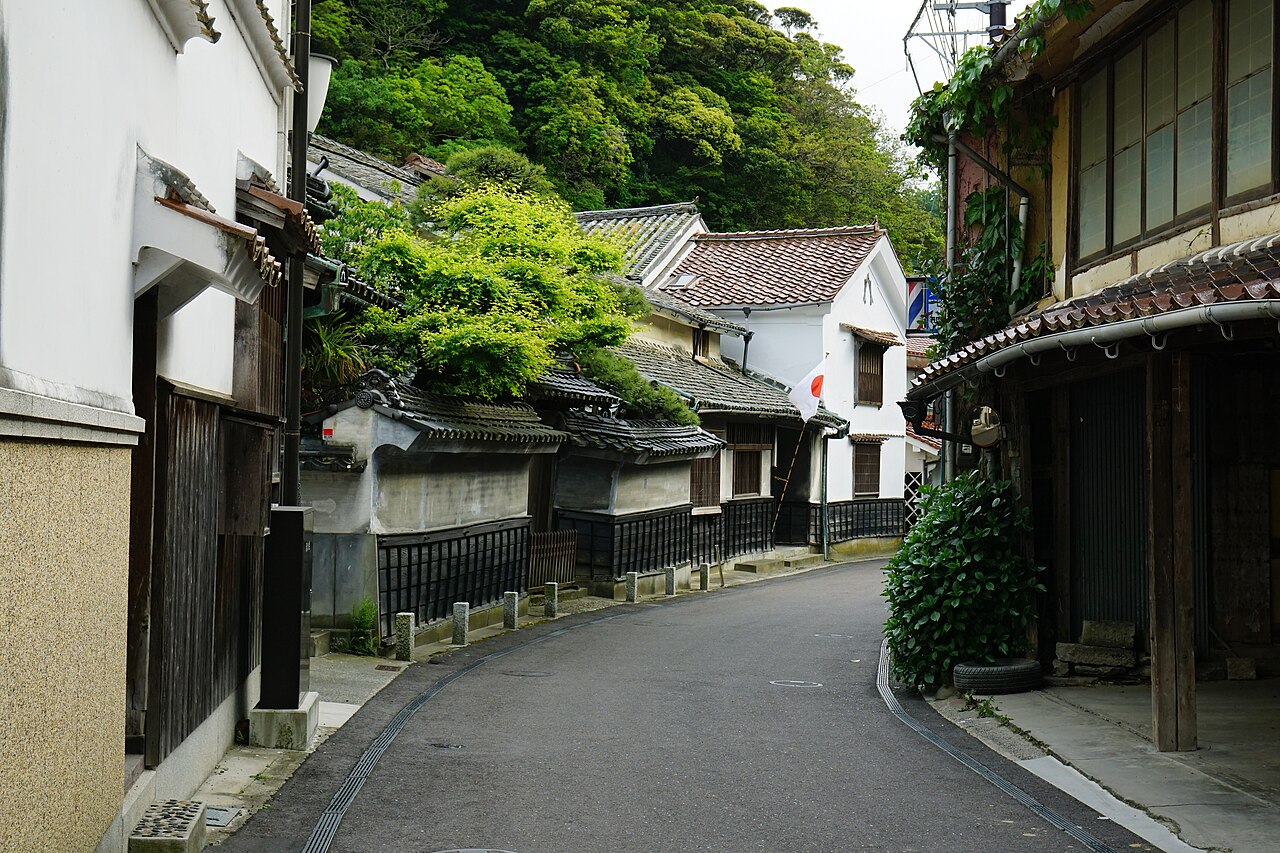 A curving lane in Yunotsu with white plastered walls, black tiled roofs and green hills behind, with a mirror on the corner.