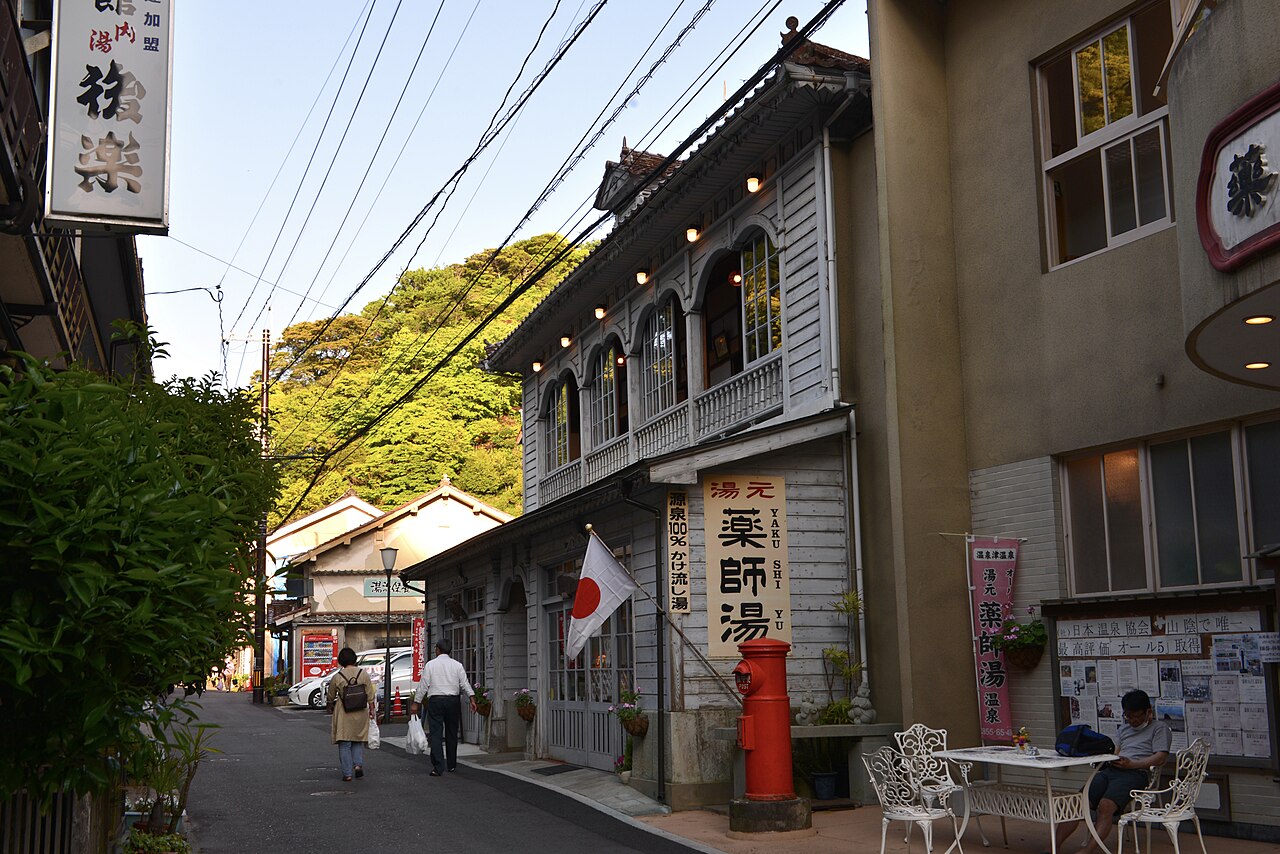 A narrow street in Yunotsu Onsen in daylight, lined with a white wooden bathhouse building with arched upper windows and an old red postbox in front.