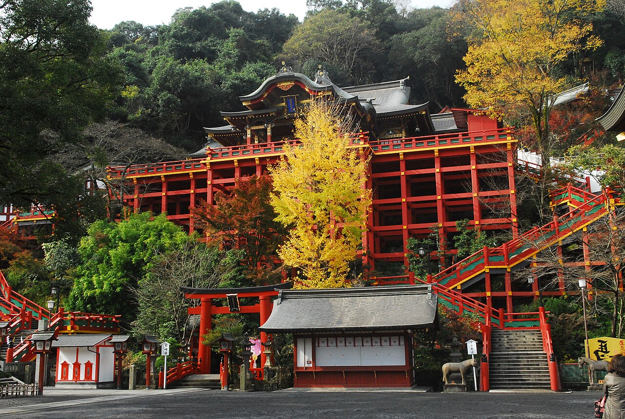 A large vermilion shrine hall stands on tall red wooden stilts against a wooded hillside, with a yellow ginkgo tree in front, red stairways climbing the slope, and a small torii and offering-stall building at the bottom.