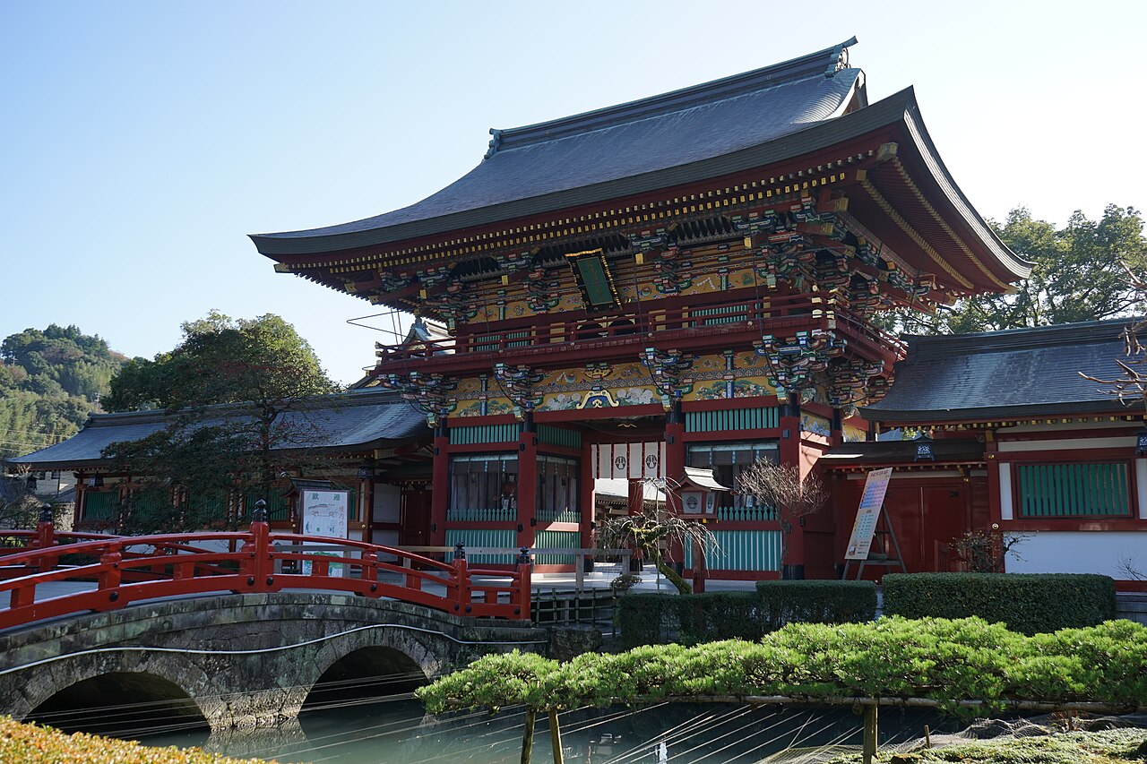 A two-storey red shrine gate with a curved grey roof and brightly painted carved brackets stands beyond a red arched bridge over a pond, under a clear blue sky.