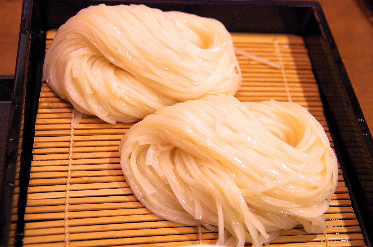 Two neat coils of thin, pale udon noodles resting on a bamboo mat in a lacquered tray.