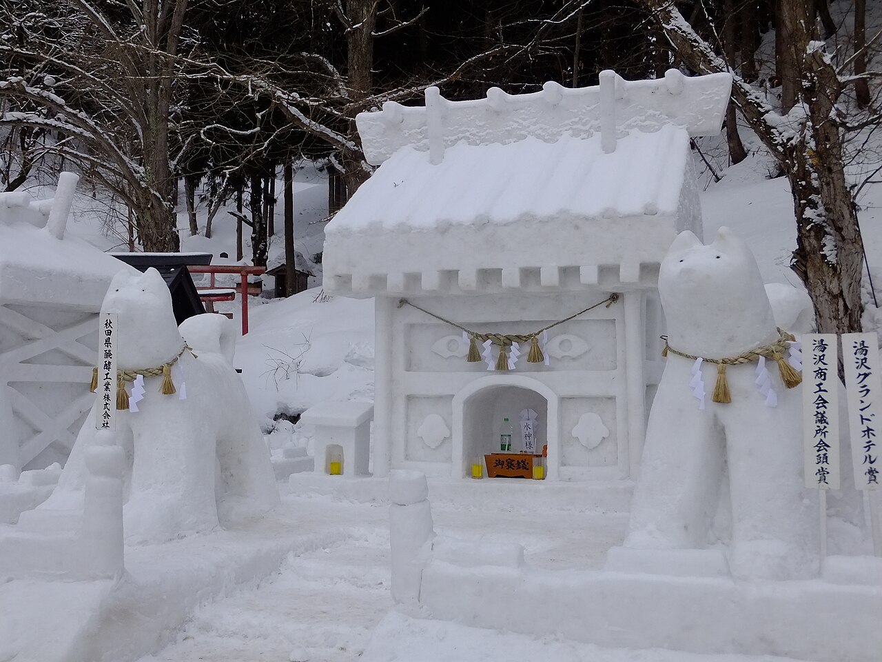 Two snow-sculpted dogs with rope collars standing on either side of a small snow shrine, with bare trees and snowy ground behind.