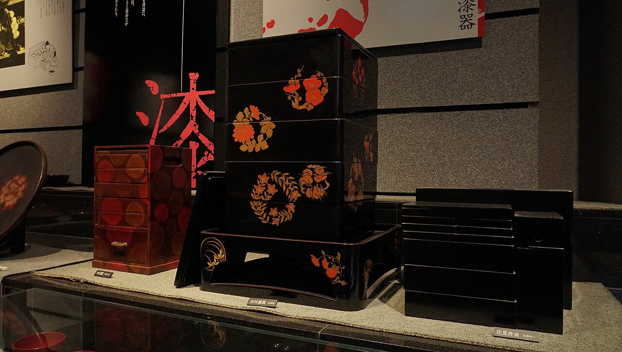 Stacked black lacquer boxes with red and gold floral designs displayed in a dim museum case.