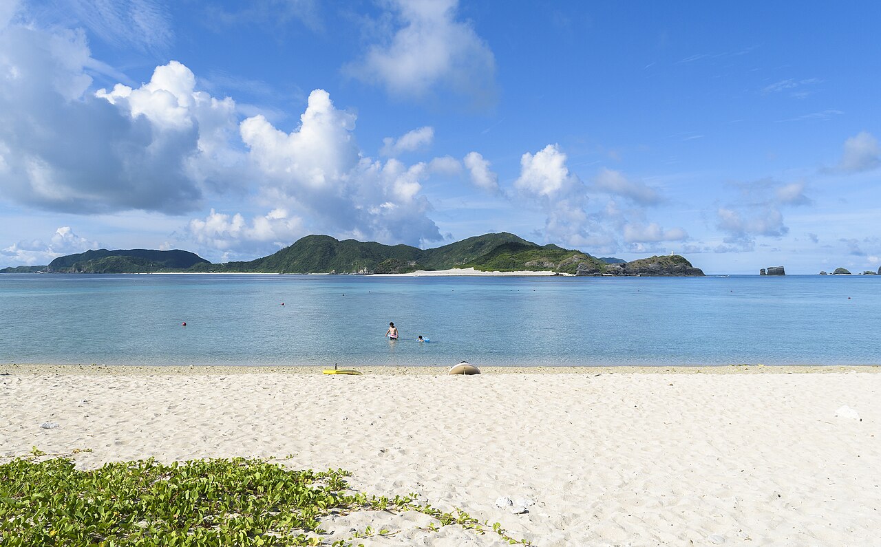 Ama Beach on Zamami Island, a wide sandy shore with calm blue water and low green islands across the strait.