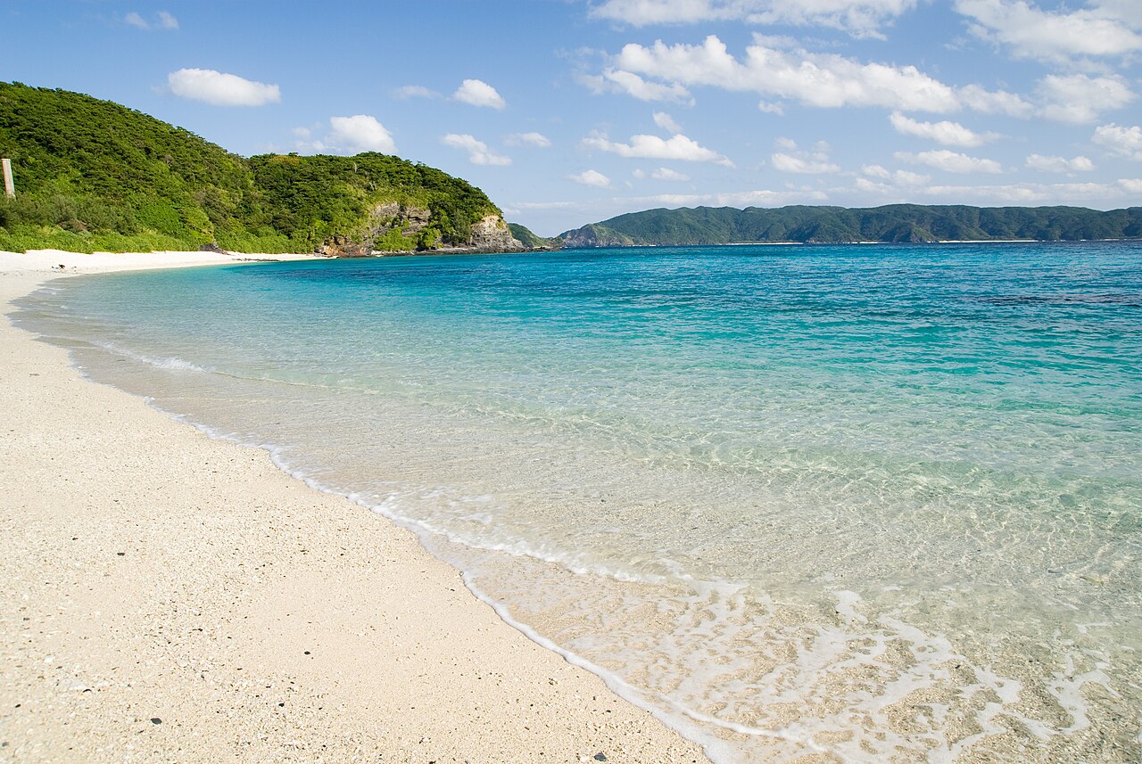 A long white sand beach with clear turquoise shallows curving toward a green headland on Zamami Island.