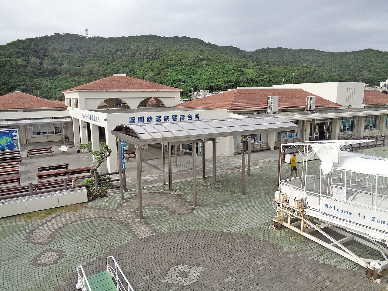 The passenger waiting terminal at Zamami port with a red-tiled roof, a covered walkway and forested hills behind.
