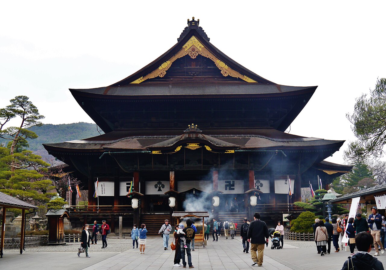 Front of the large Zenko-ji main hall with its tall layered roofs, seen across the stone-paved courtyard on an overcast day