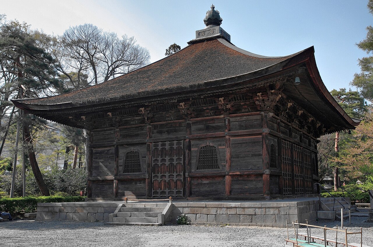 Square wooden Kyozo sutra library standing on a low stone base, with a dark tiled roof topped by a metal finial and pine trees around it