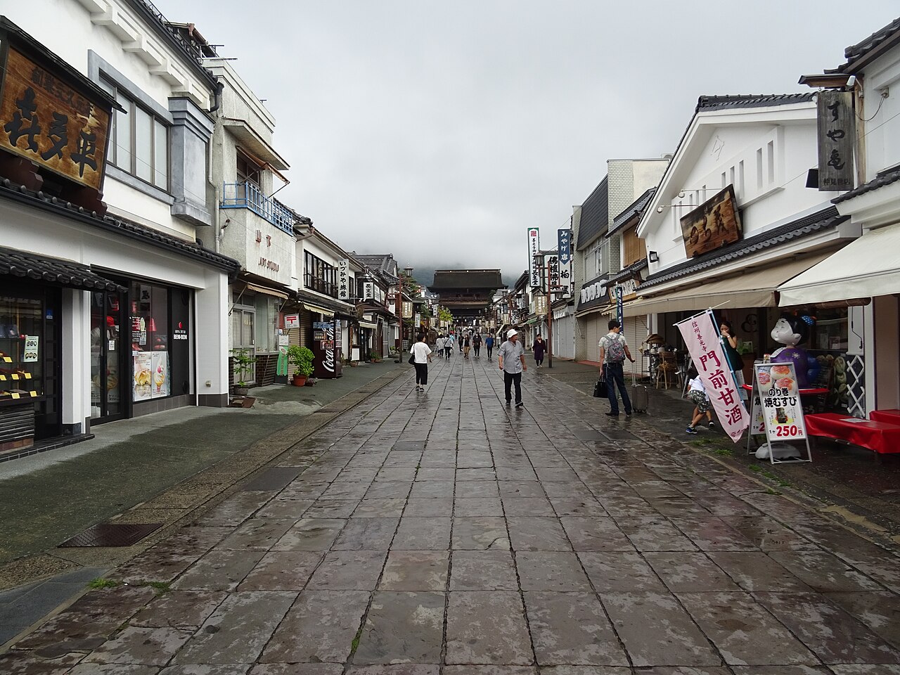 Pedestrians walking up the stone-paved approach lined with shops and paper banners, with a large temple building and green hills ahead
