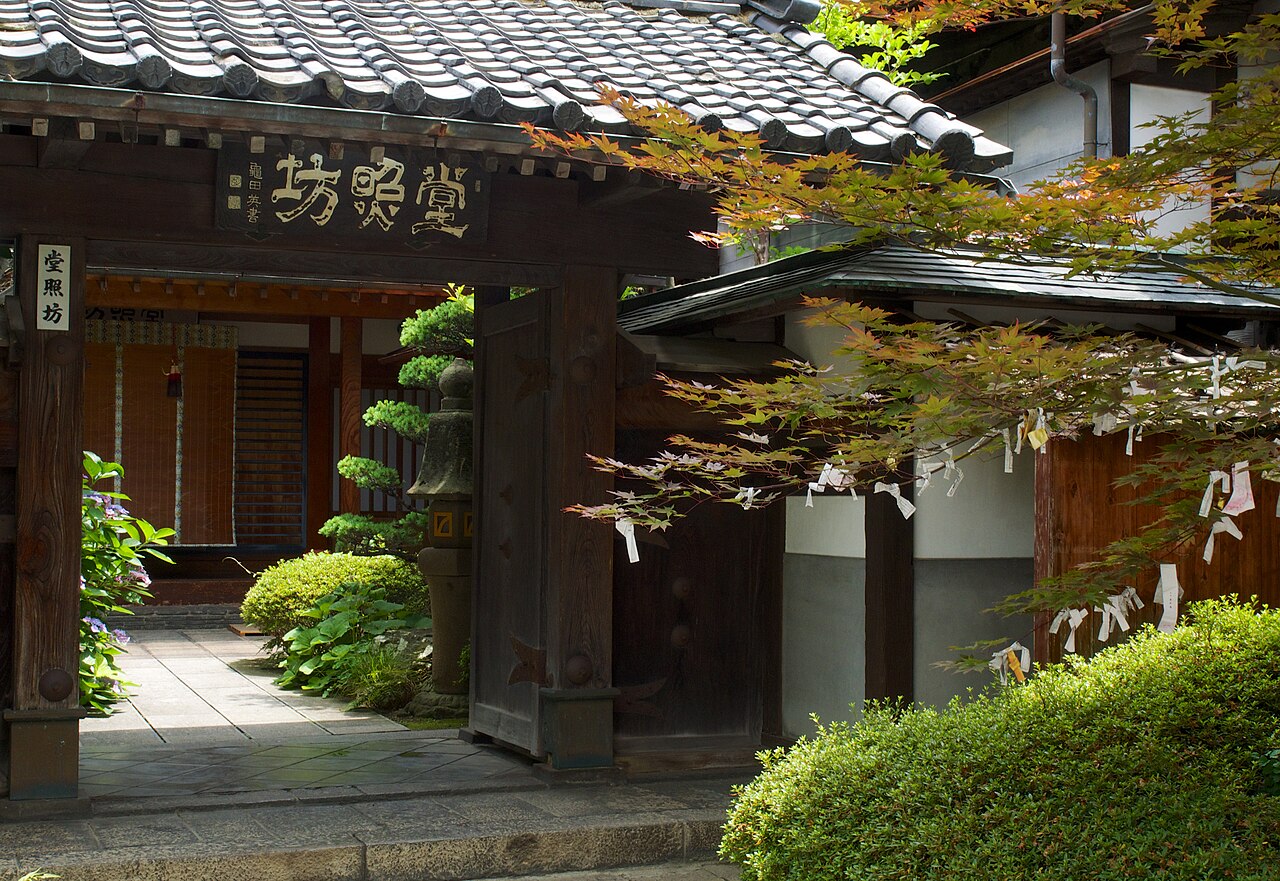 Wooden gate with a hanging signboard and tiled roof, opening onto a small stone-paved garden with a stone lantern and trimmed shrubs