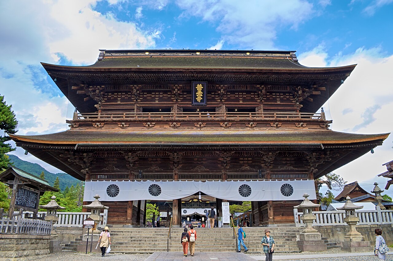 The two-story Sanmon gate of Zenko-ji with its dark wooden brackets and white curtain, seen from the foot of the stone steps under a blue sky