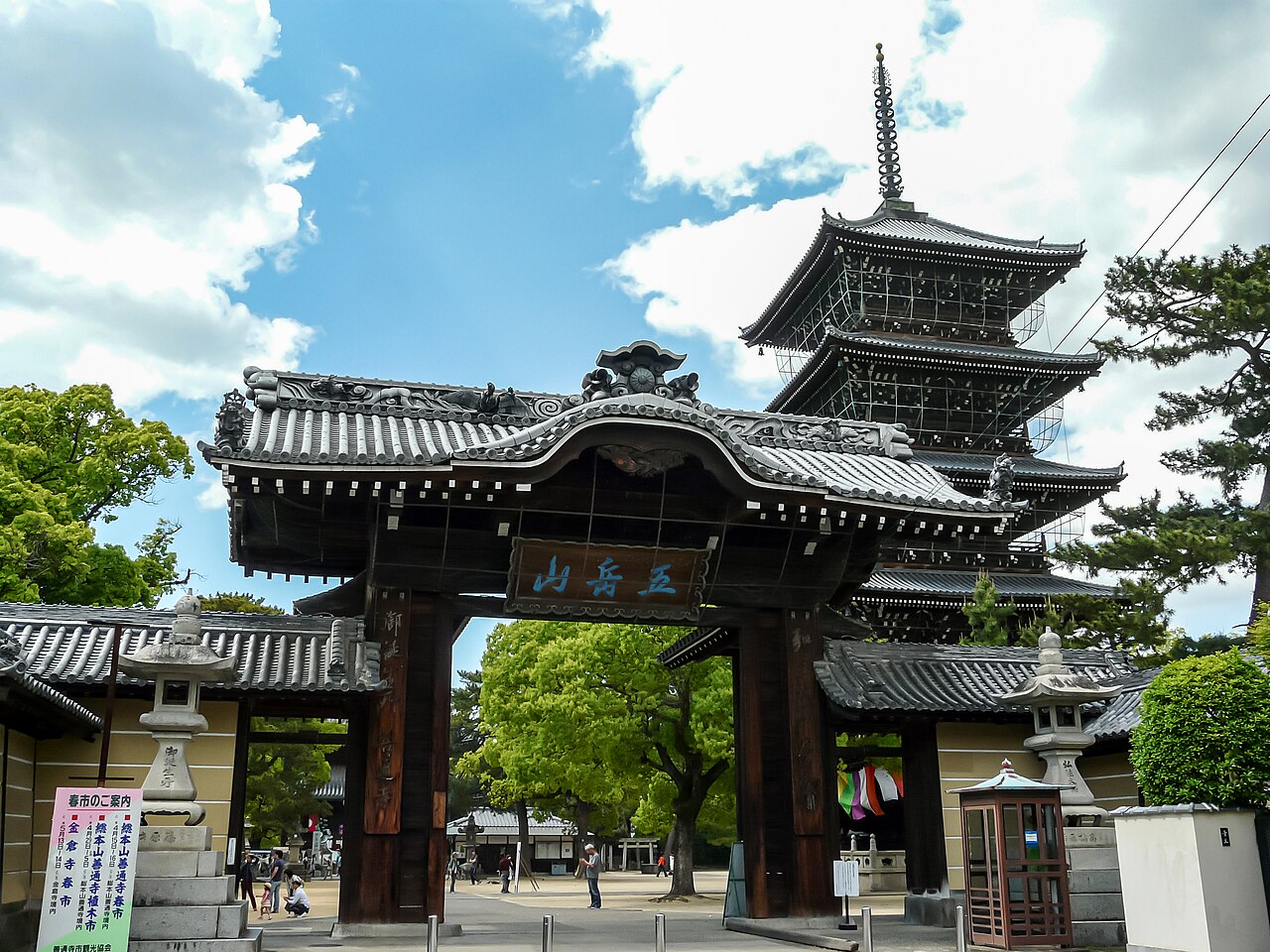 The ornate south gate of Zentsu-ji temple seen across a paved approach, with the tiers of the five-storied pagoda rising behind it under a blue sky.