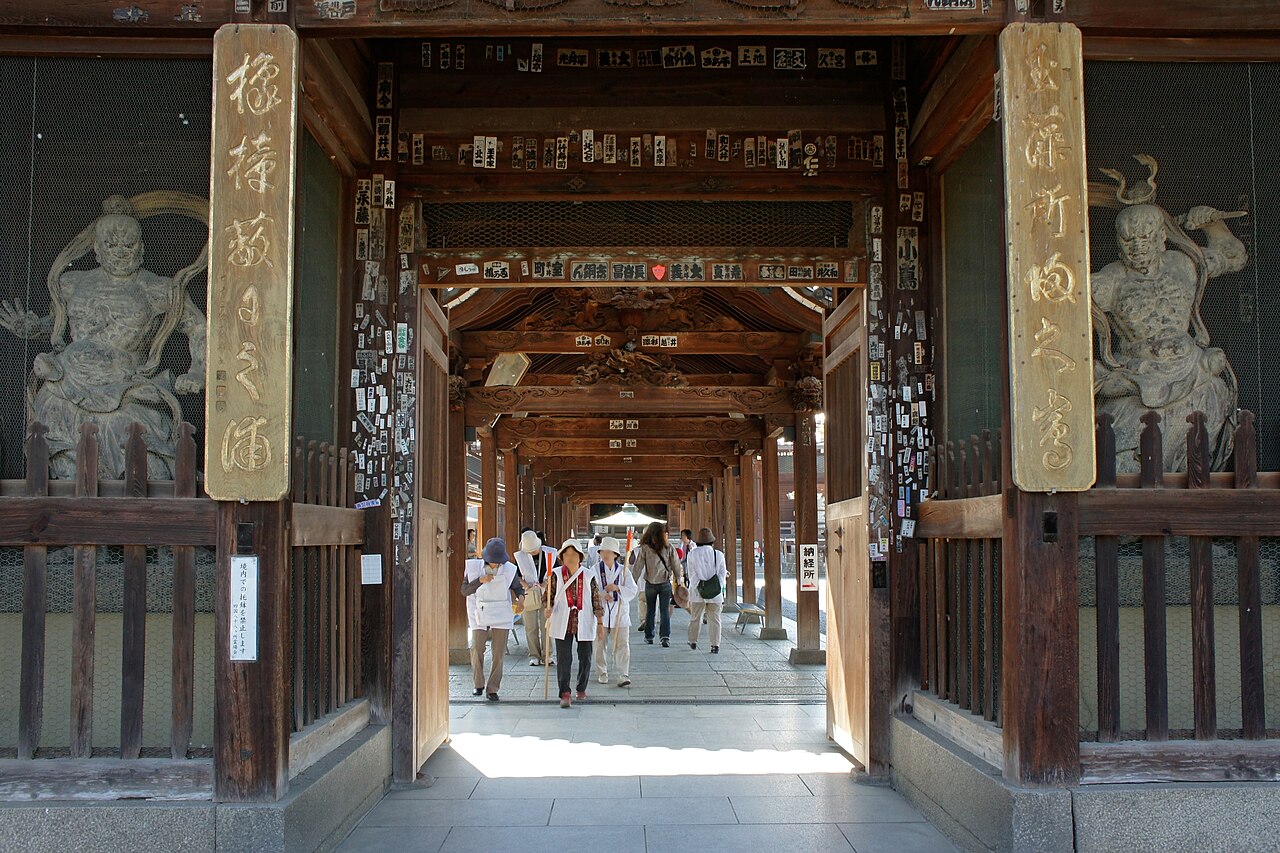 Looking through a wooden temple gate with two guardian statues in its side bays, down a long roofed corridor where a few visitors walk.