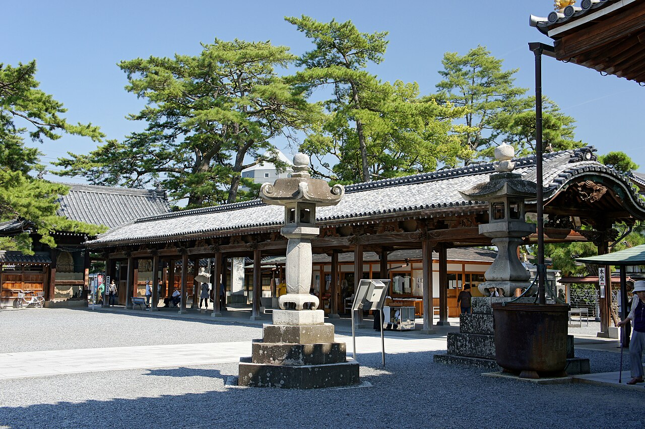 A wide gravel temple precinct with tall stone lanterns in the foreground and a long tiled-roof wooden hall under pine trees on a sunny day.