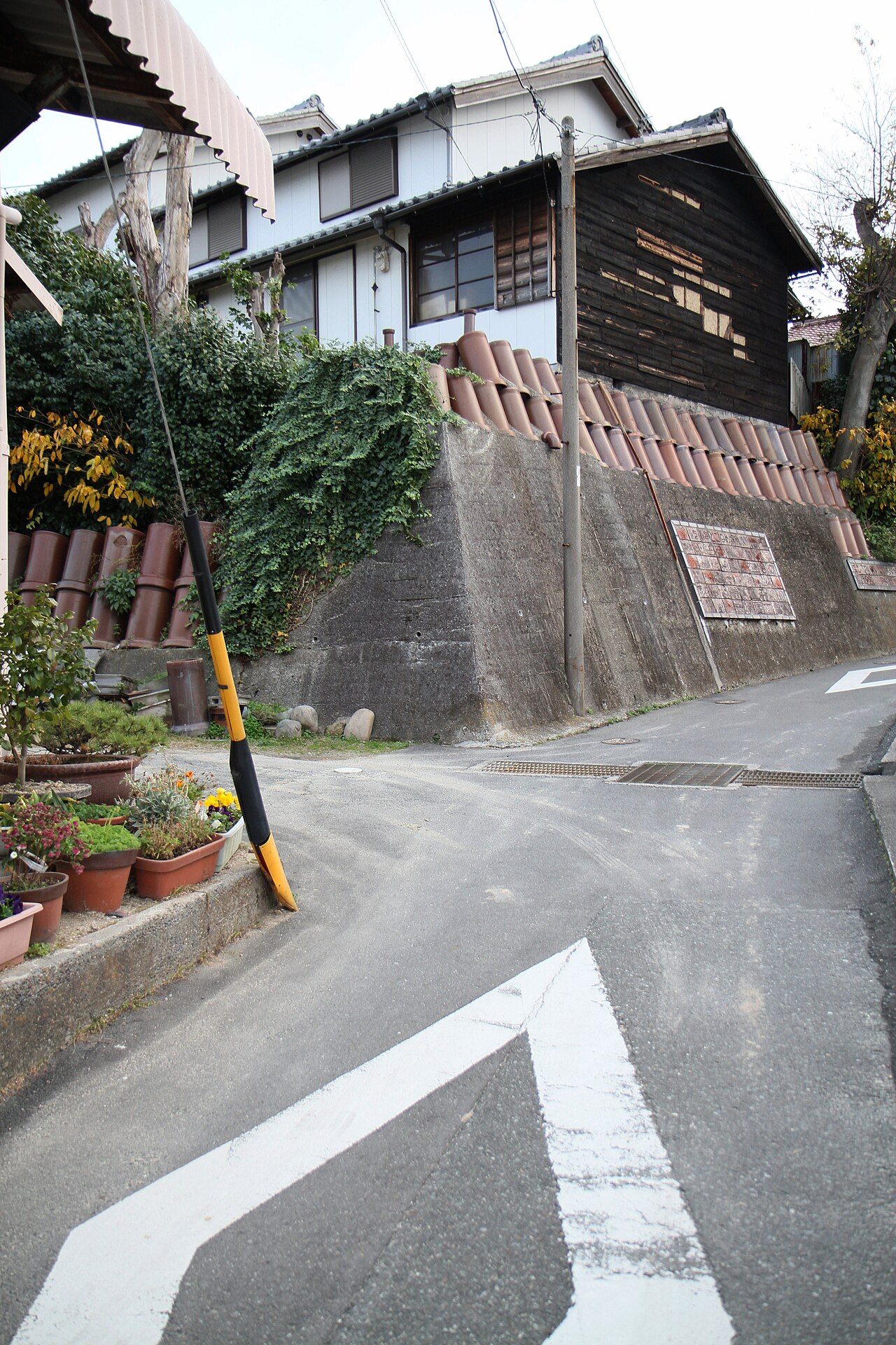 A retaining wall on the Tokoname pottery footpath built from stacked clay drainage pipes and shochu jars, with potted plants along the lane