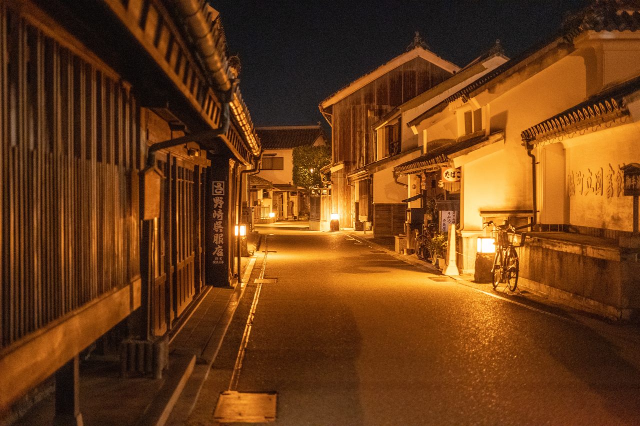 A row of whitewashed udatsu shops along a street in Wakimachi lit at night by warm lamps, with a bicycle parked outside one shopfront.