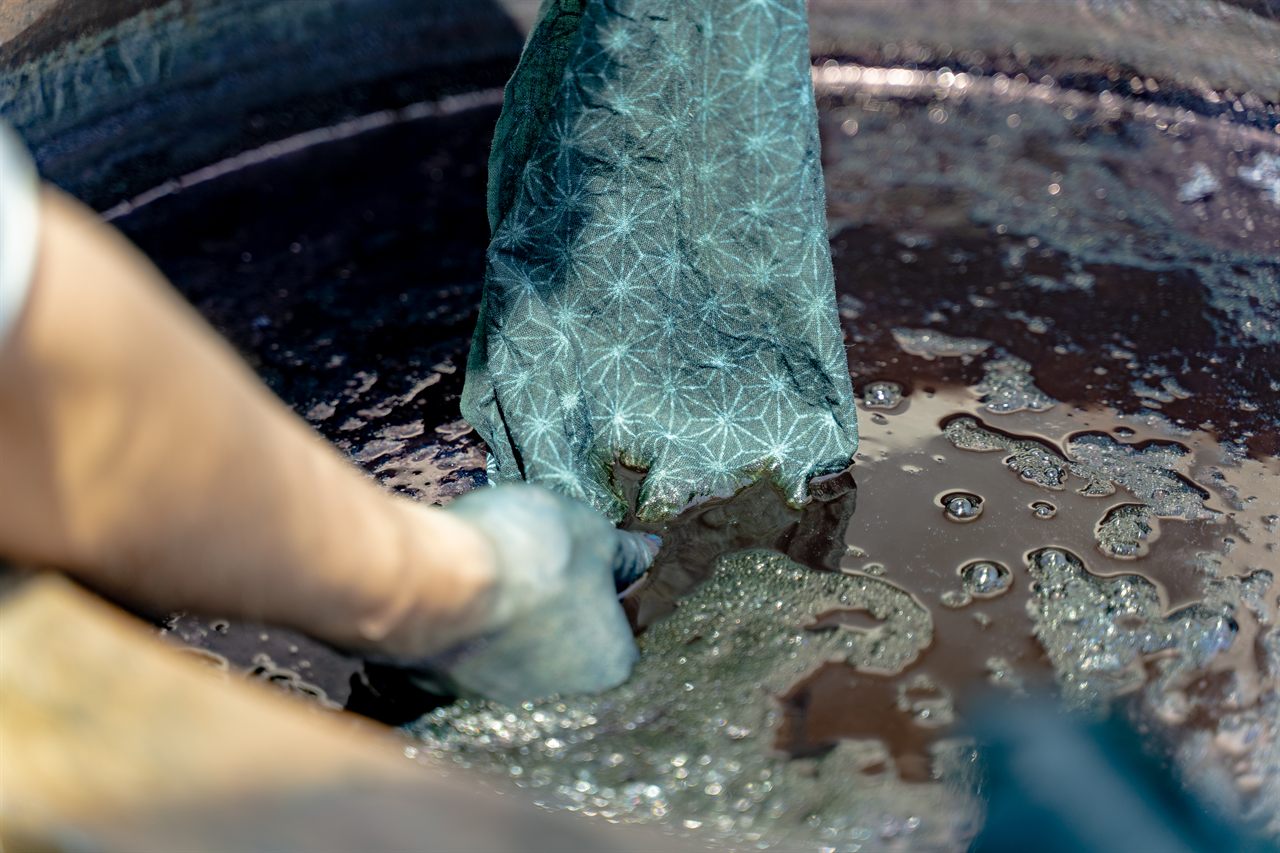 A gloved hand lifts a star-patterned cloth dyed blue and white out of a dark indigo dye vat.
