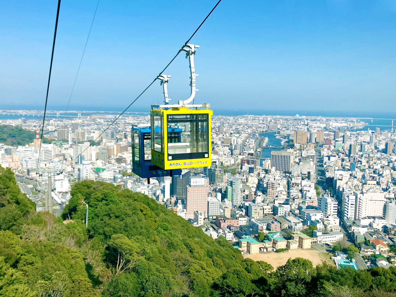 A yellow and blue ropeway gondola descending over green hillside with the dense city of Tokushima, a river and the sea stretching out below under a clear blue sky