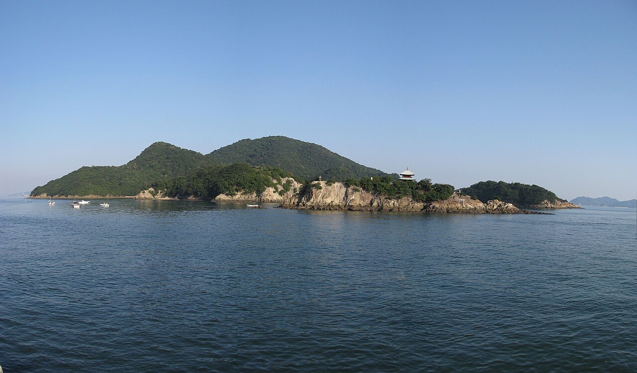 Sensuijima's forested twin hills rise above a rocky point in the Seto Inland Sea, a small pavilion with a peaked roof standing on the rocks and a few fishing boats resting on the calm water below.